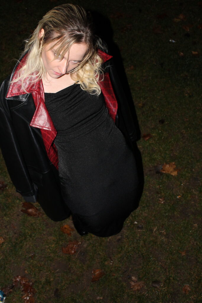 “Moody nighttime portrait shot from above of a person in a black dress and black jacket with a red collar, standing on grass with fallen leaves, looking downward.”